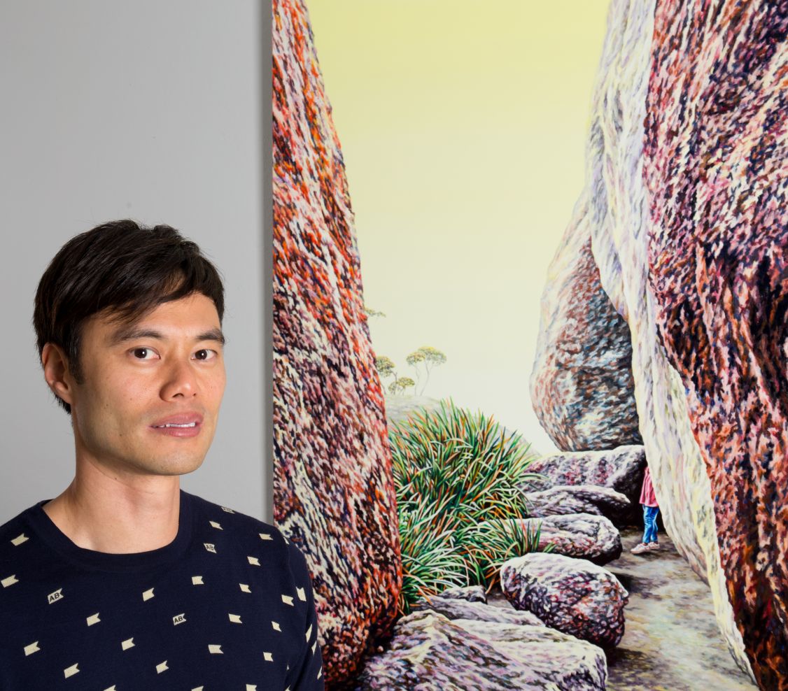 Man smiling standing in front of a painting with large rocks, bushes, trees and a person standing behind a rock