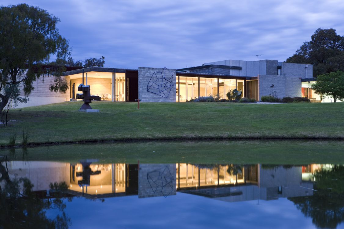 Picture of McClelland Gallery building on top of a grassy hill, with a lake in the foreground. The lake depicts the reflection of the building