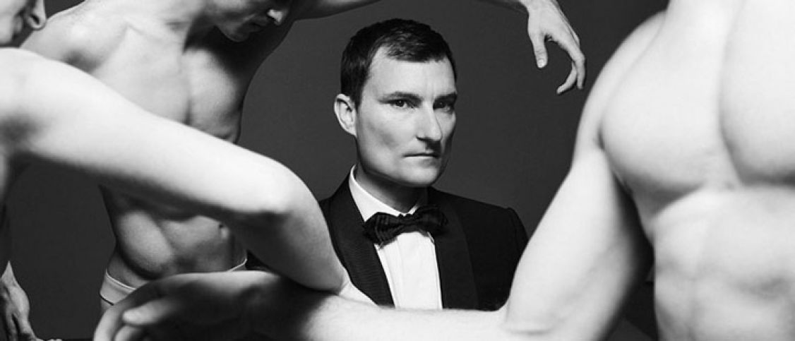 Black and white image of man in a bow tie and suit staring at the camera with naked torsos around him