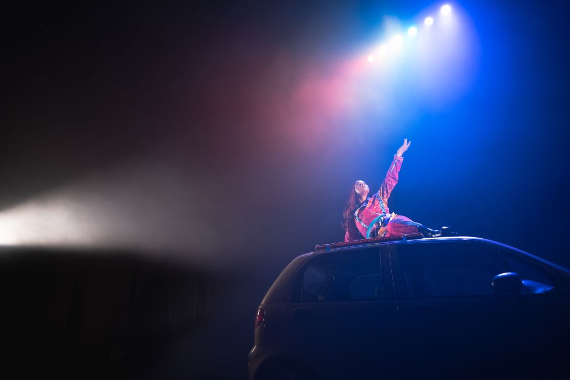 A woman with long hair laying on top of a car with her arm reaching up into the air